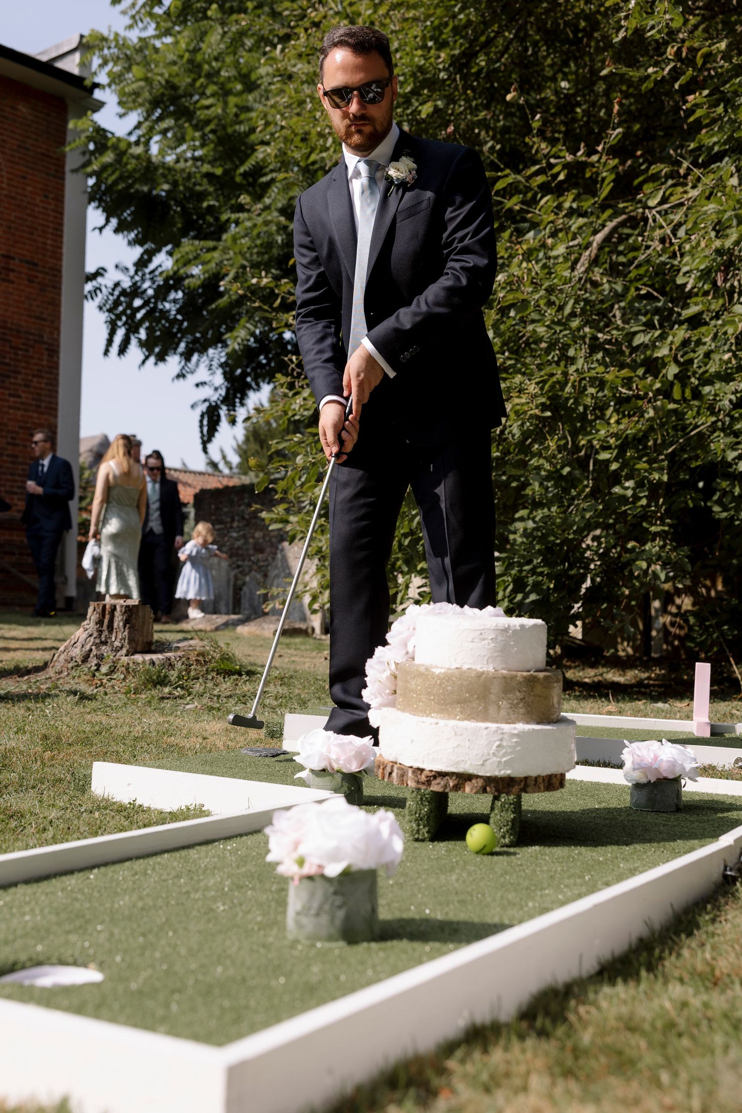 Groom in suit playing mini golf near a tiered wedding cake on a green course at an outdoor wedding reception.