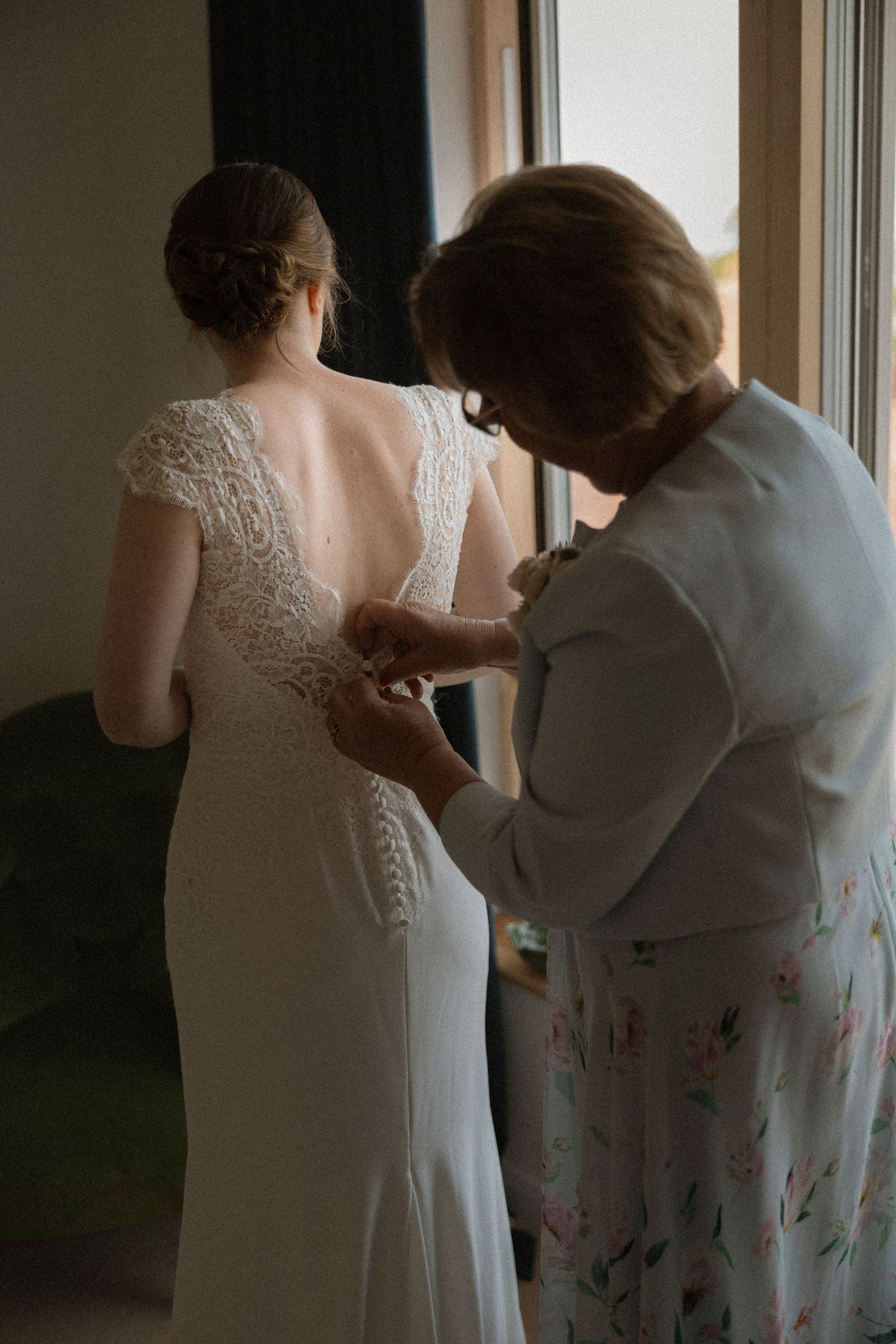 Mother helping bride button up elegant lace wedding dress near window before ceremony.