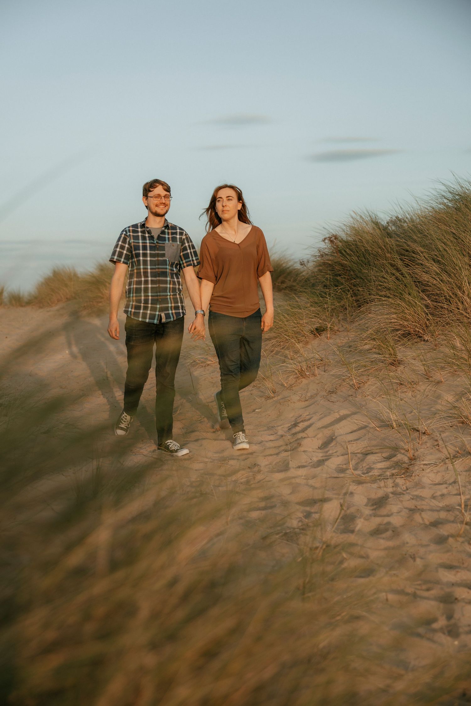Couple walking hand in hand along sandy beach dunes at dusk.