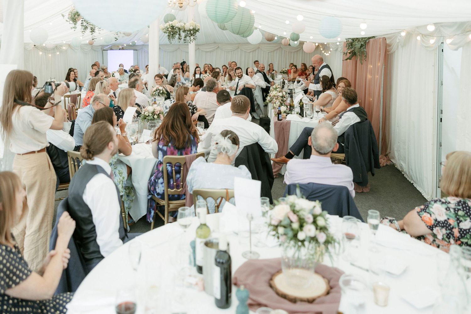 Elegant wedding reception in a marquee with guests seated at round tables adorned with floral centerpieces and soft decor.