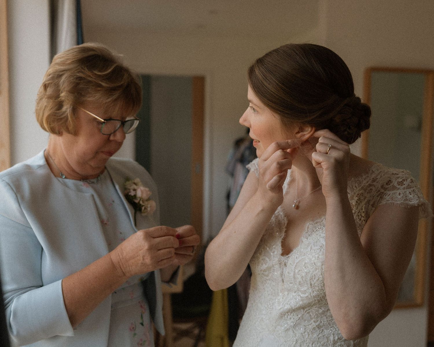 Bride in lace wedding gown adjusts earring while older woman in blue jacket assists during wedding preparations.