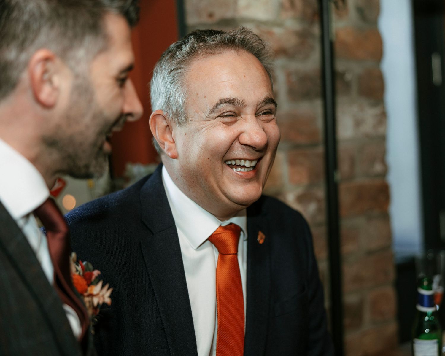 Two guests sharing a joyful moment at a wedding reception against exposed brick wall.