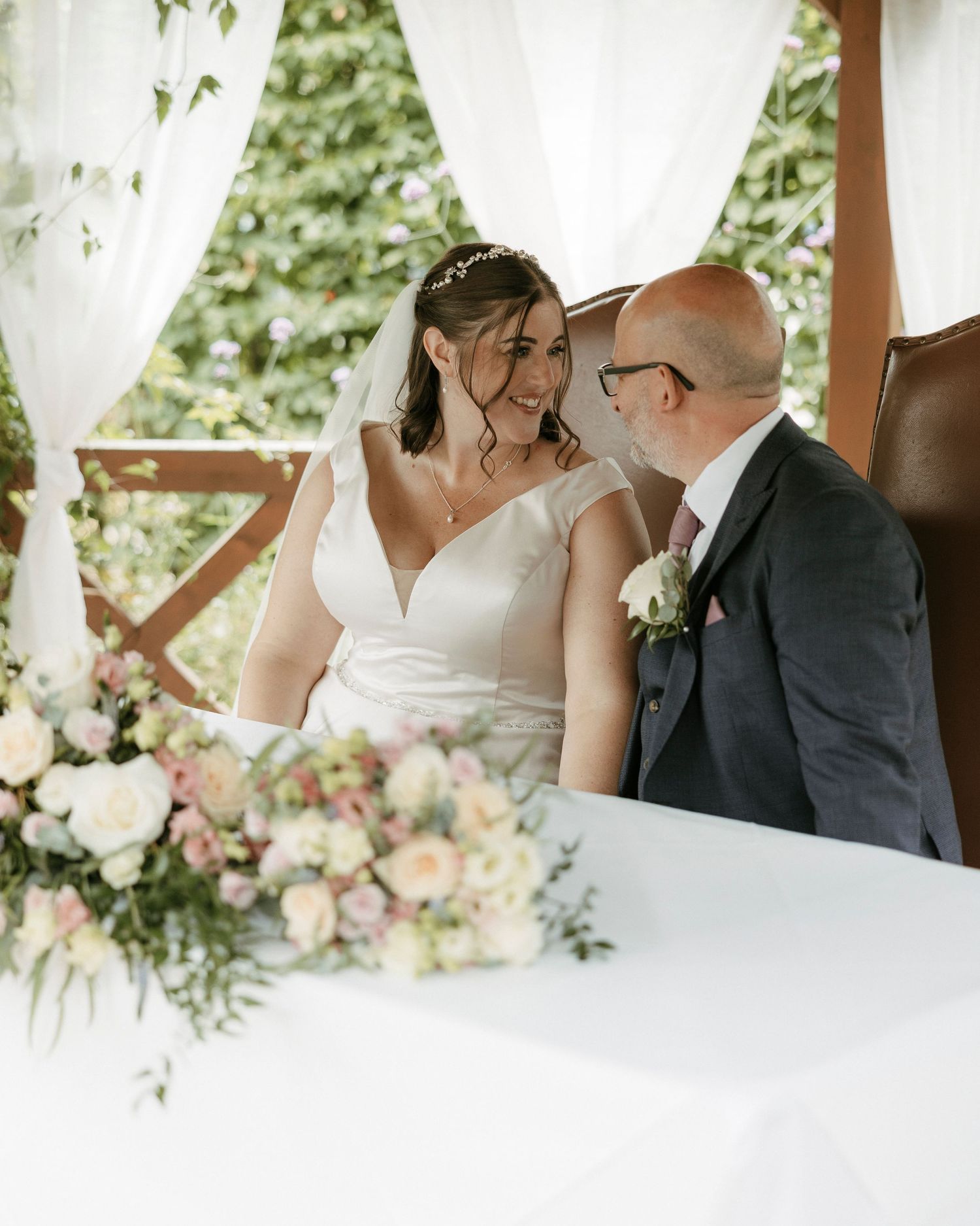 Bride and groom share a joyful moment at wedding ceremony table adorned with pastel floral arrangements.