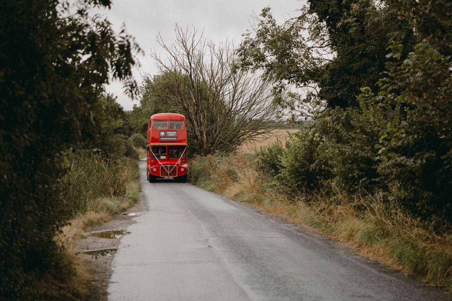 A classic red double-decker bus travels down a narrow rural road lined with lush green trees.