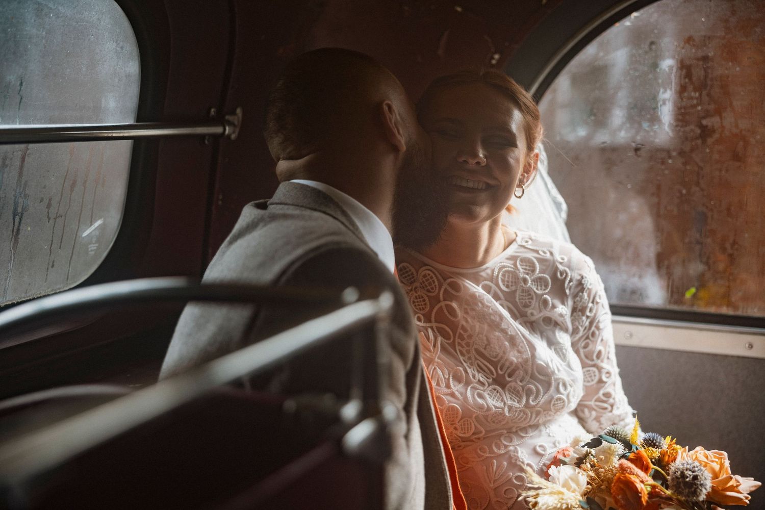 Bride in lace wedding dress holding bouquet sits beside groom in vintage car on wedding day.