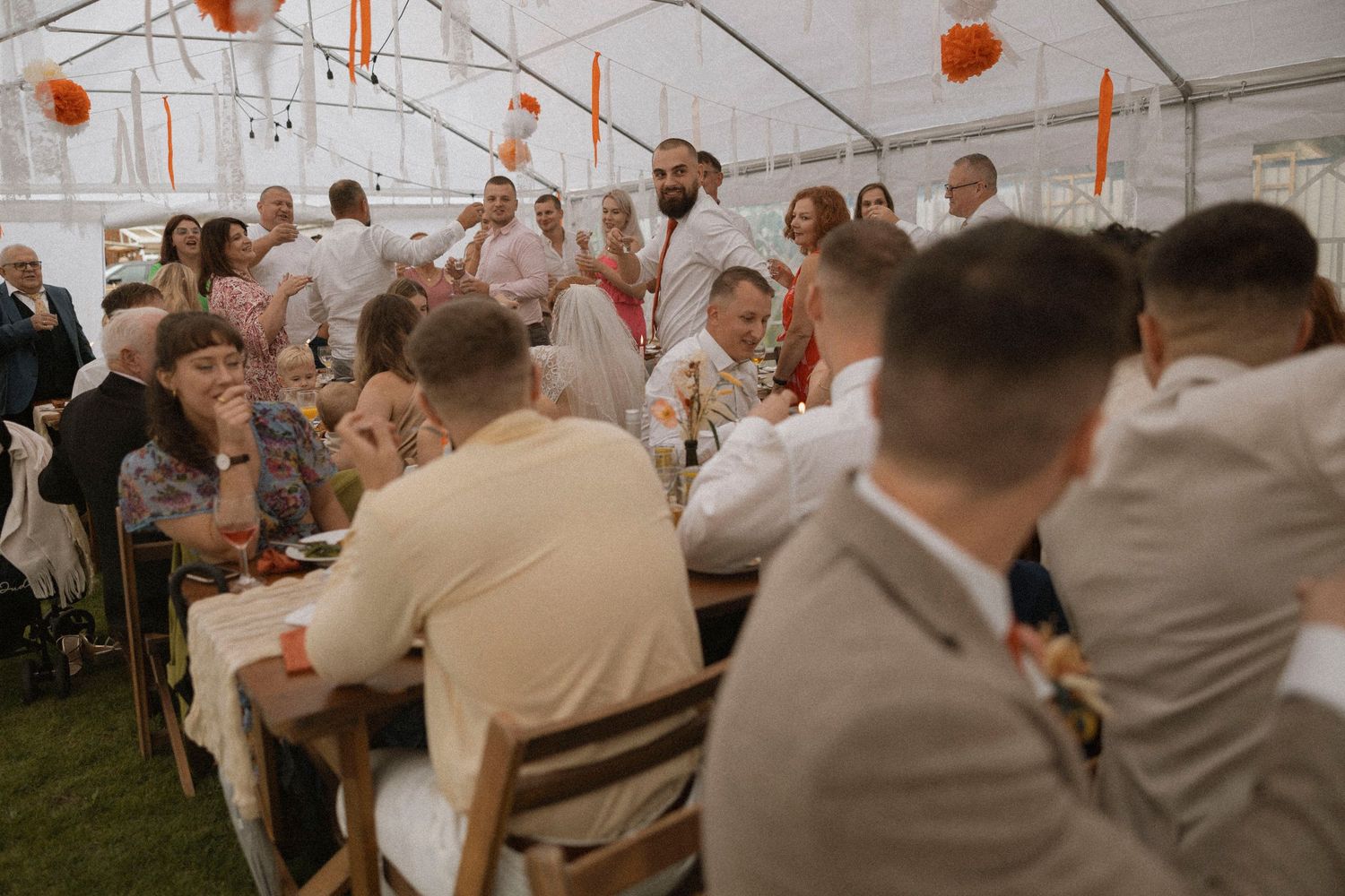 Guests seated at long tables during a wedding reception inside a decorated marquee tent with orange accents.
