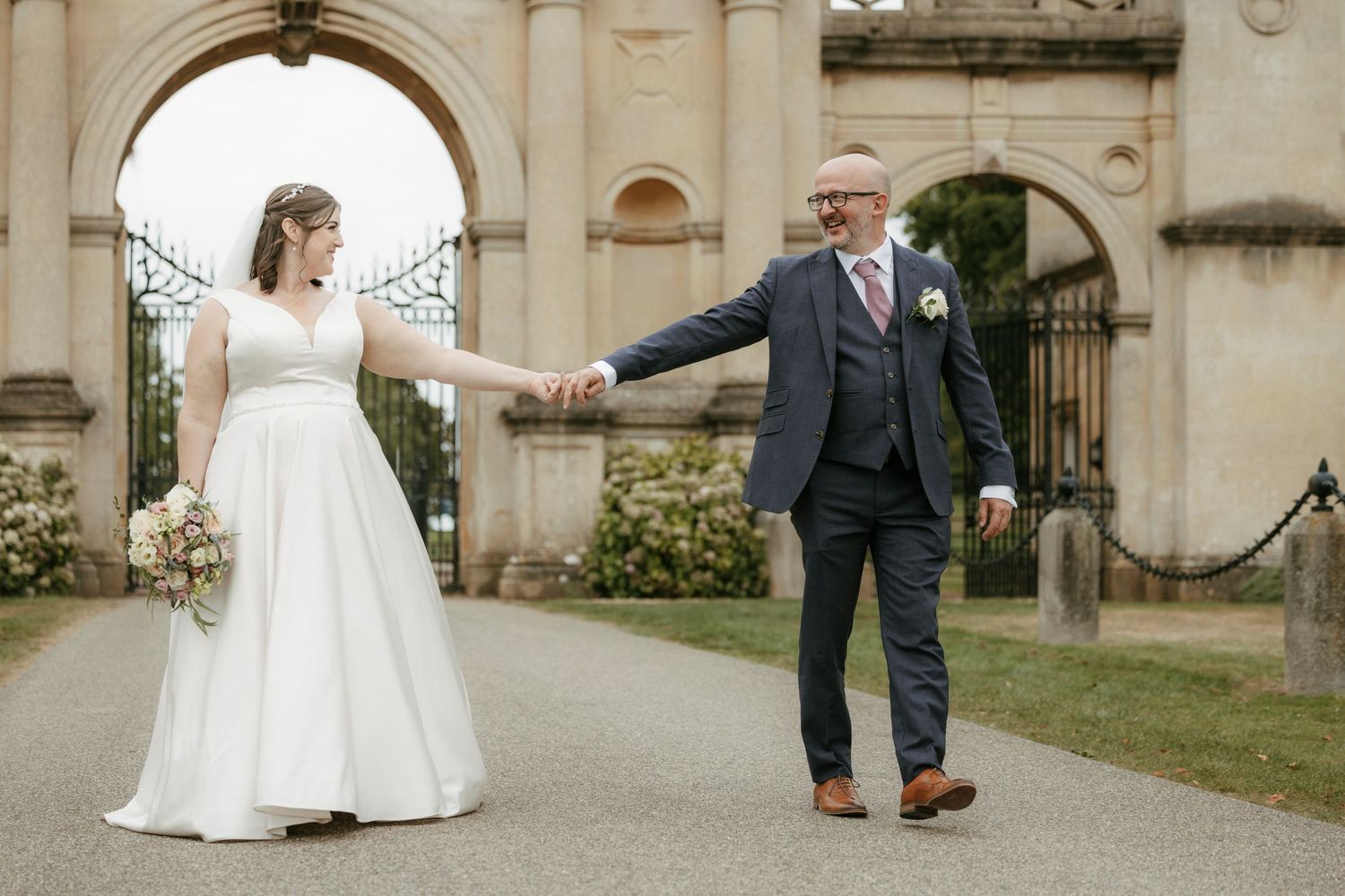 Bride and groom hold hands walking through ornate stone gates at an elegant outdoor wedding venue.