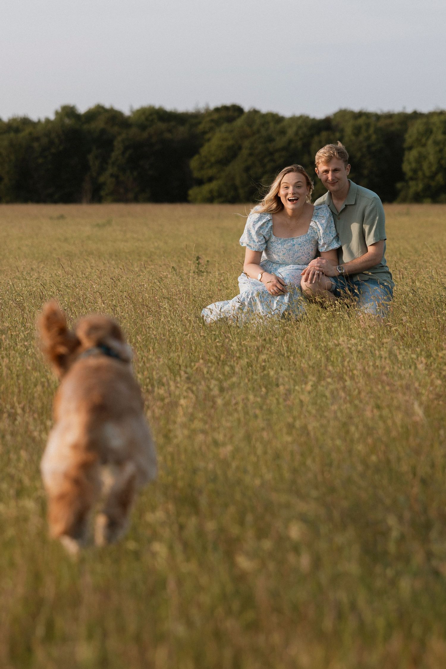 A happy couple sits in a green field laughing as their golden retriever dog runs toward them.