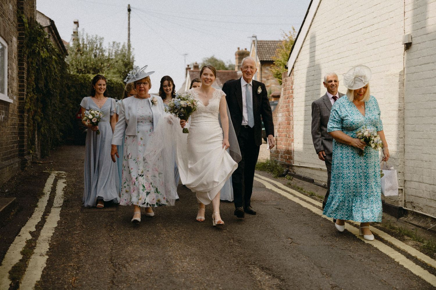 Bride walks with wedding party down a charming village street, bridesmaids in blue dresses and guests in elegant attire.