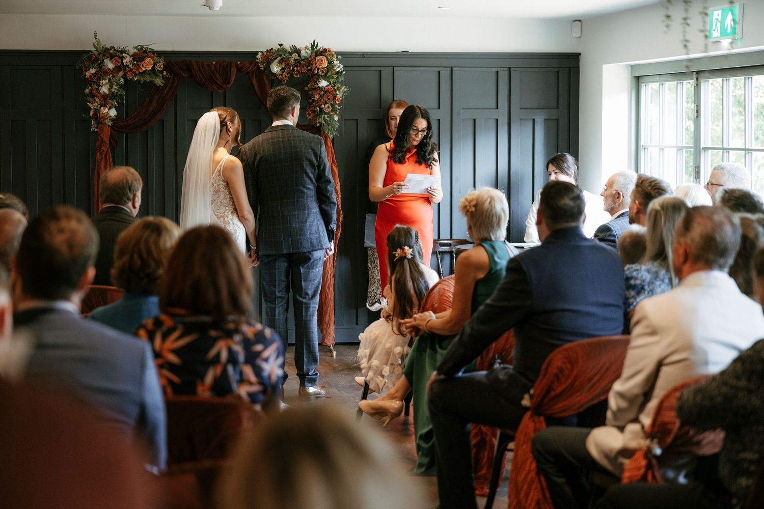 Wedding ceremony with bride and groom at altar while officiant in red dress addresses seated guests.