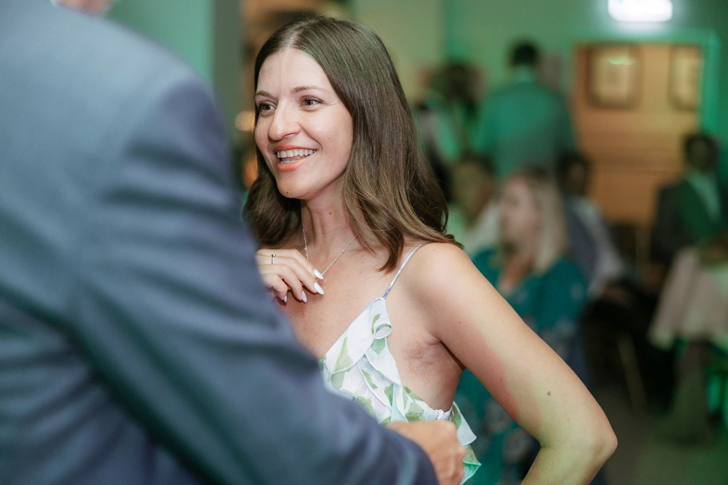 A smiling woman in a floral dress at an indoor event, looking over her shoulder in a warmly lit room.