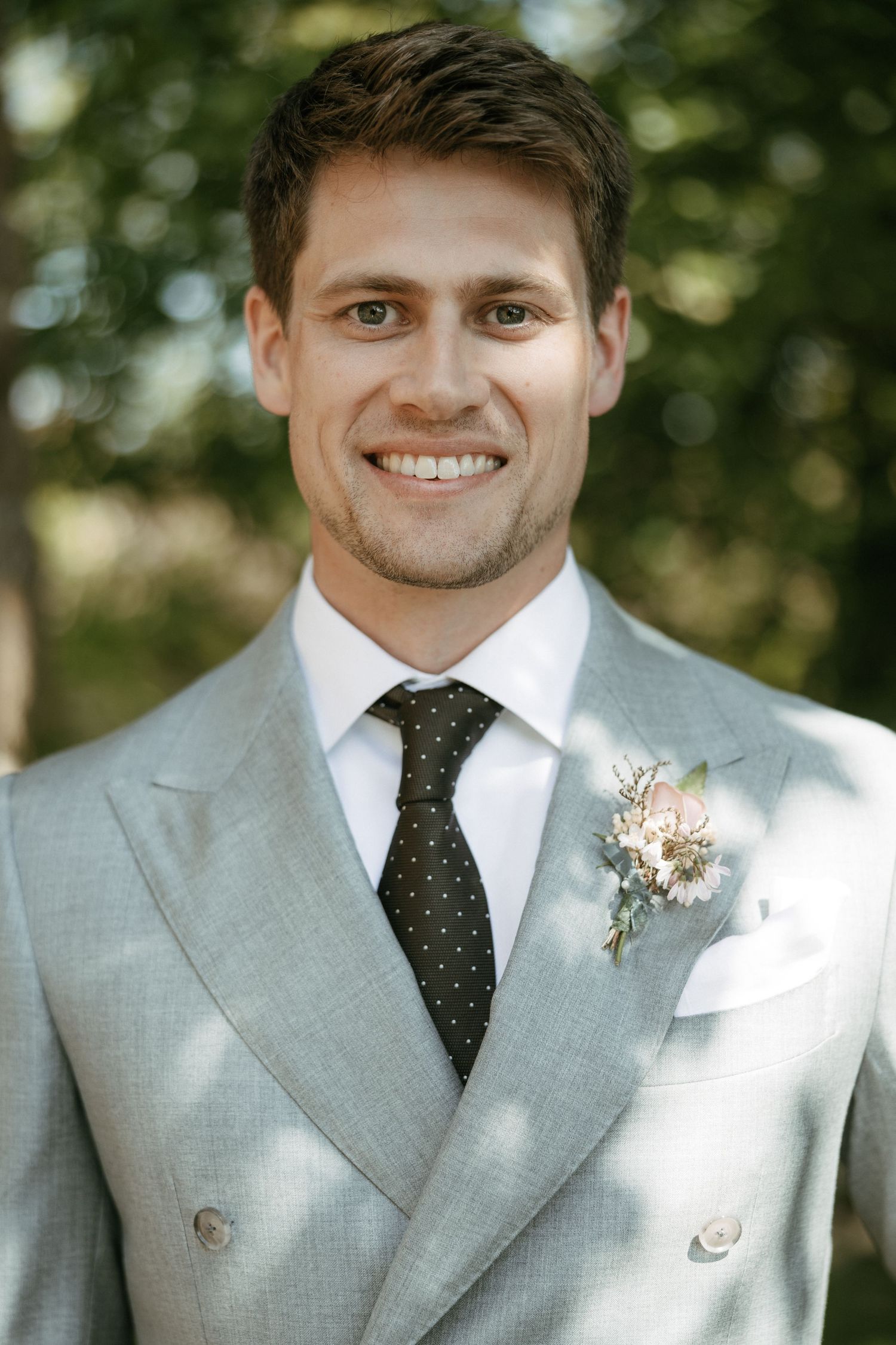 Smiling groom in light gray double-breasted suit with dark polka dot tie and floral boutonniere outdoors.