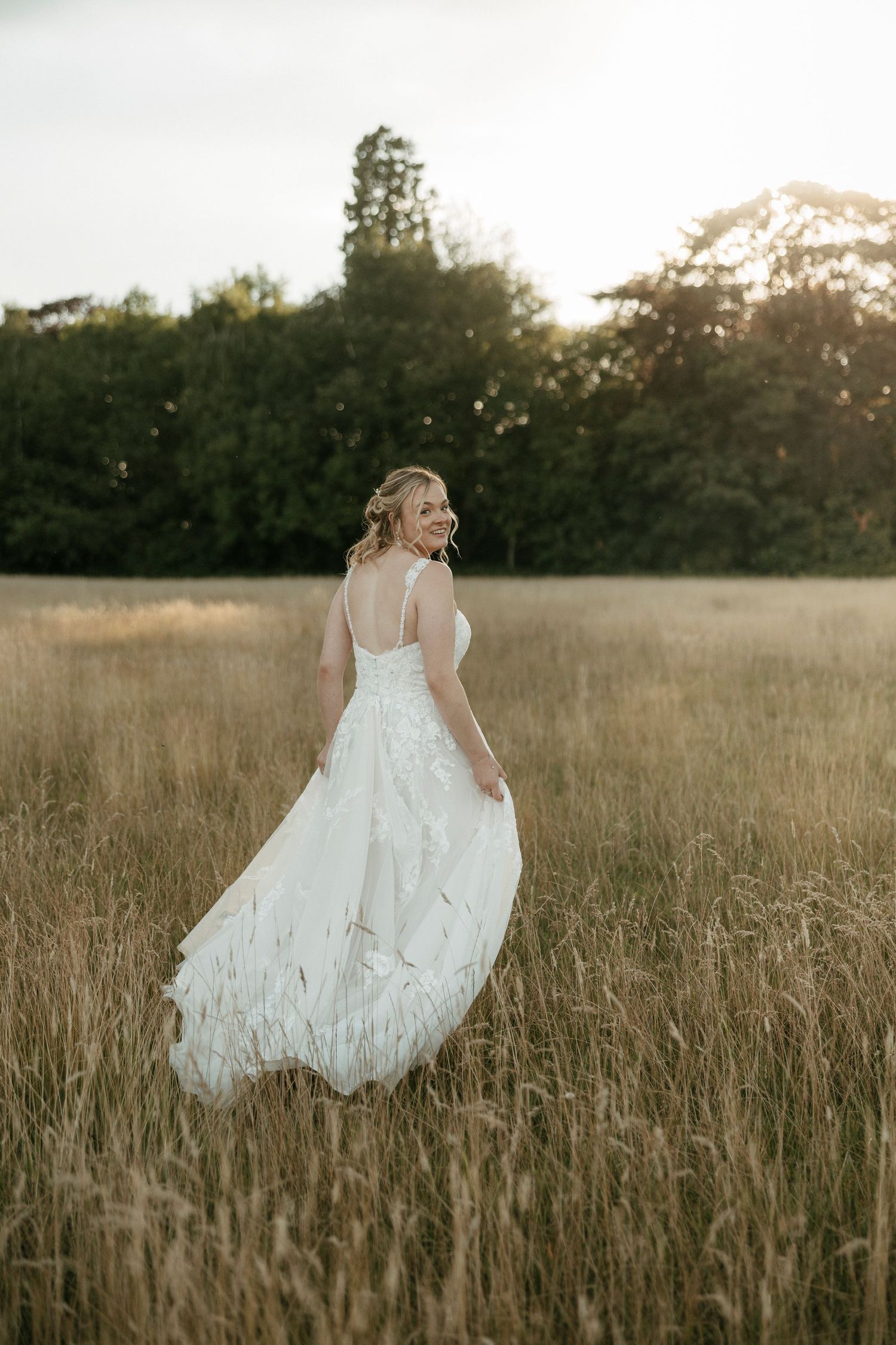 A flowing white wedding dress catches the golden light as someone stands in a serene meadow at sunset.