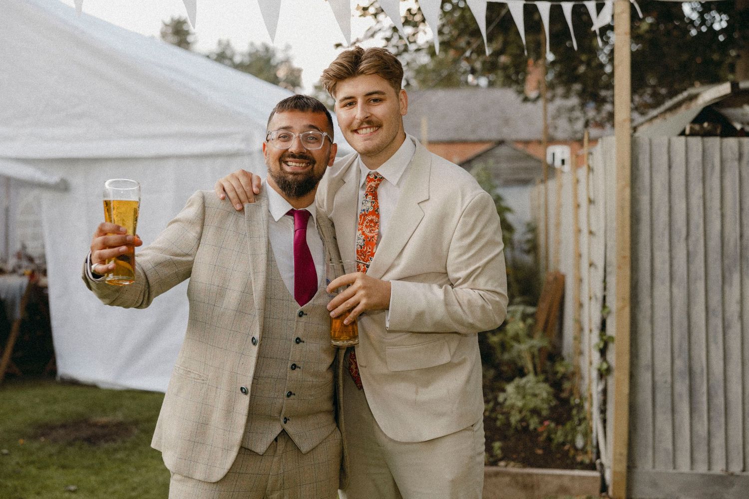 Two men in beige suits holding drinks and smiling at an outdoor wedding reception celebration.