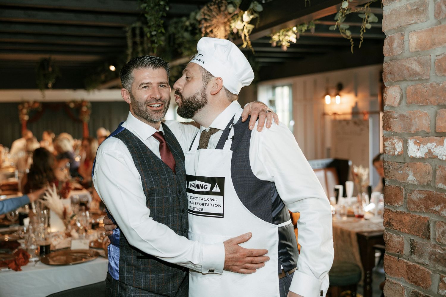 Two men in formal attire embrace warmly at a wedding reception, one wearing a chef hat and apron.