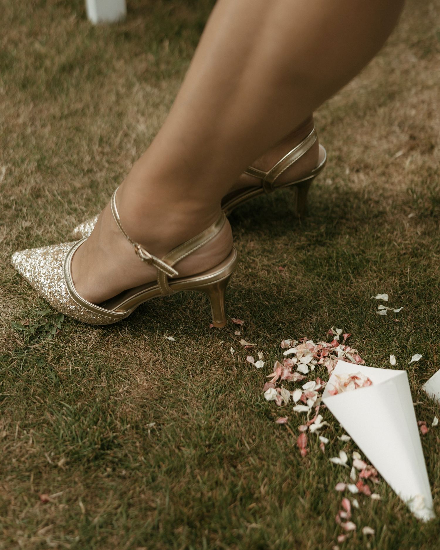 Gold glitter kitten heel shoes with ankle strap on grass with floral confetti cone at a wedding.