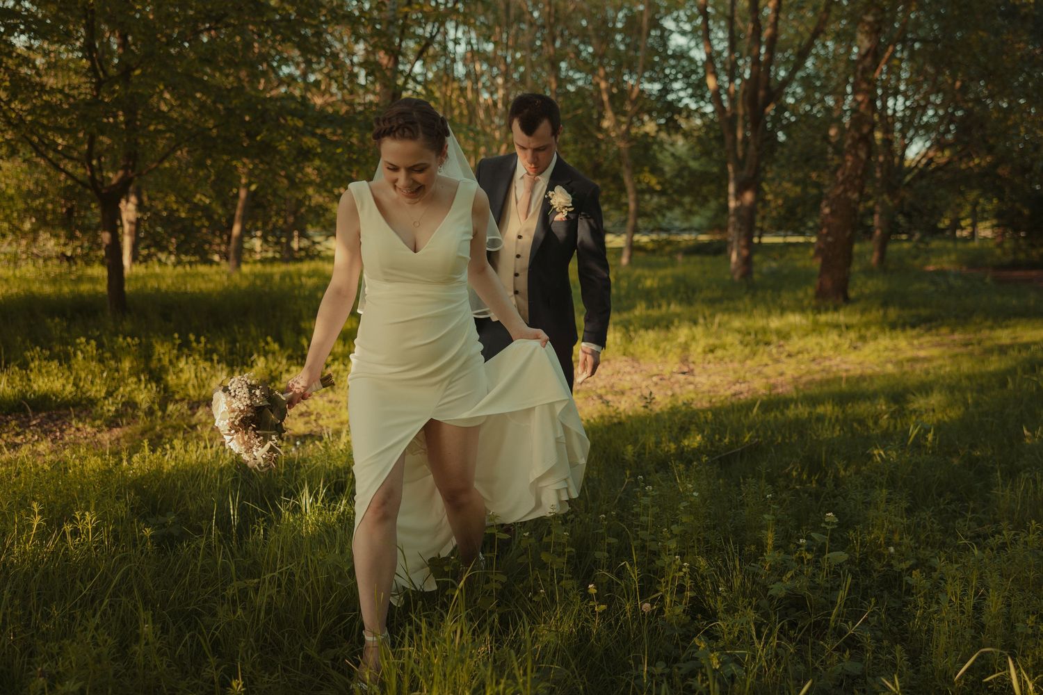 Bride and groom walk hand-in-hand through lush green meadow with trees, bride holding bouquet in golden sunlight.