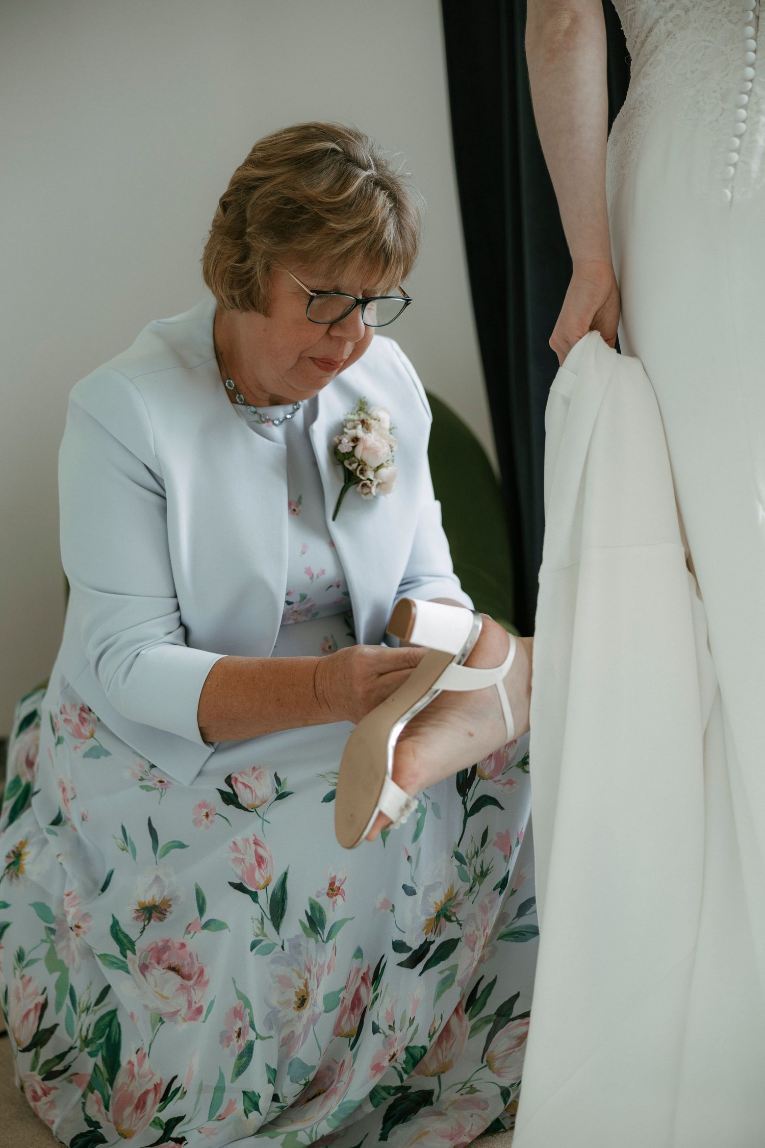 Mother of the bride helping bride put on wedding shoes, wearing floral dress and corsage.