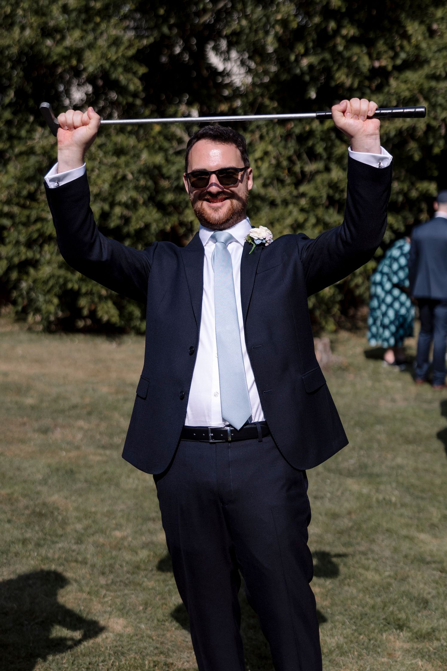Groom in navy suit and sunglasses triumphantly raises a cane overhead outdoors at a wedding reception.