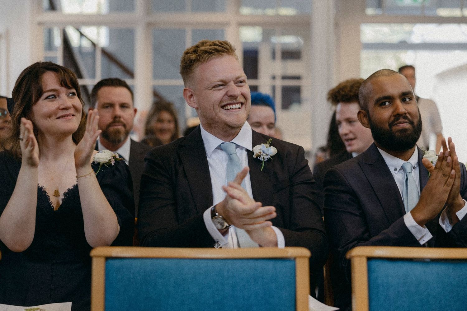 Three guests clapping and smiling joyfully during a wedding ceremony seated in blue chairs.