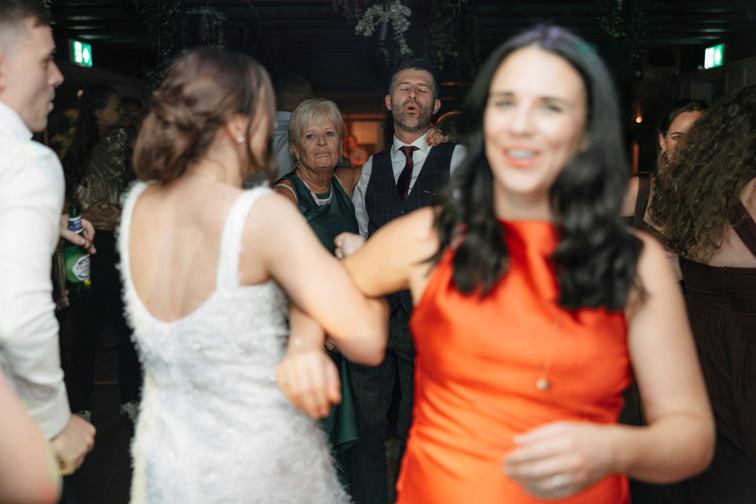 Wedding guests dancing at a lively indoor reception, bride in white gown and woman in orange dress in foreground.