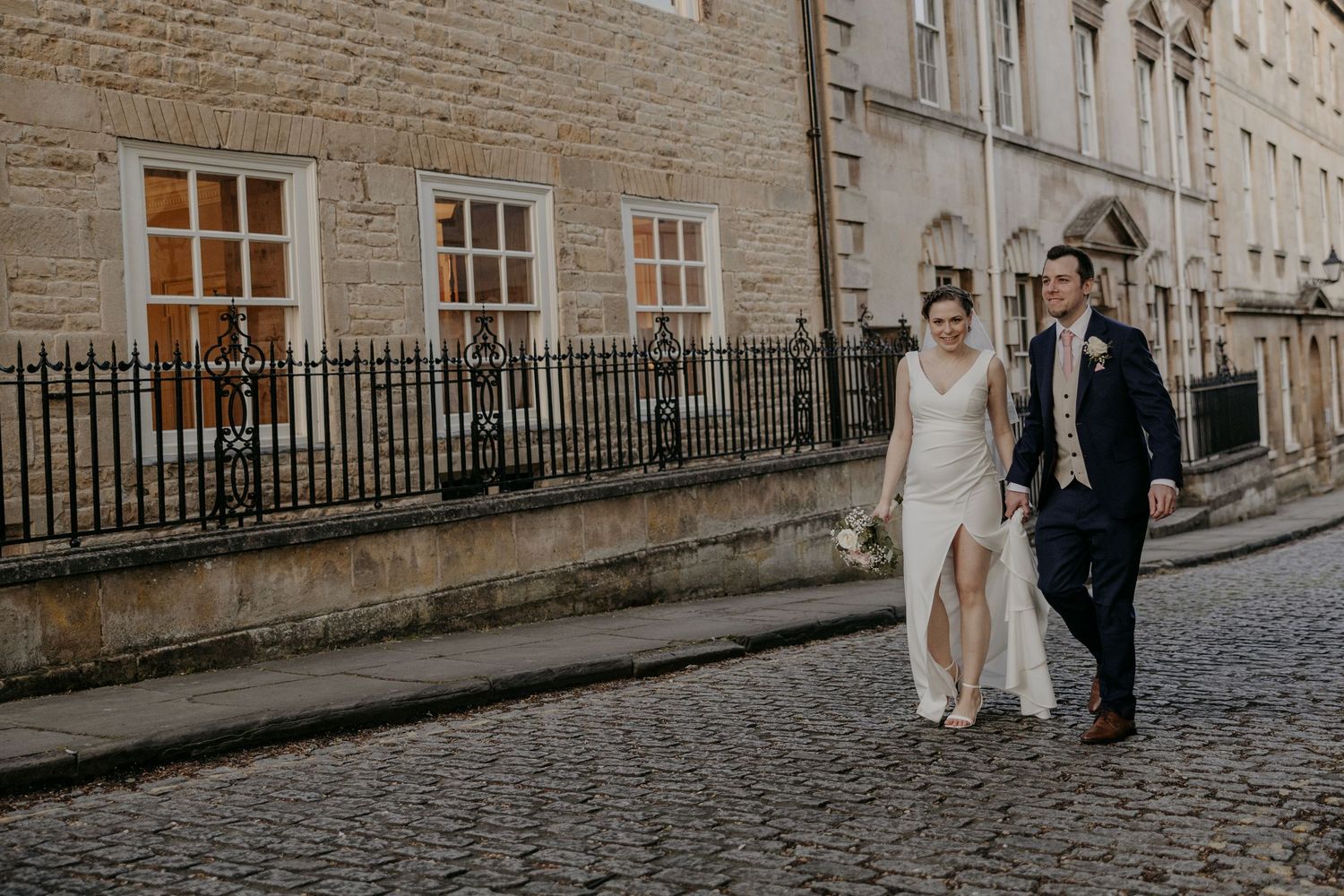 Bride and groom walking hand in hand on cobblestone street beside historic stone building with iron railings.