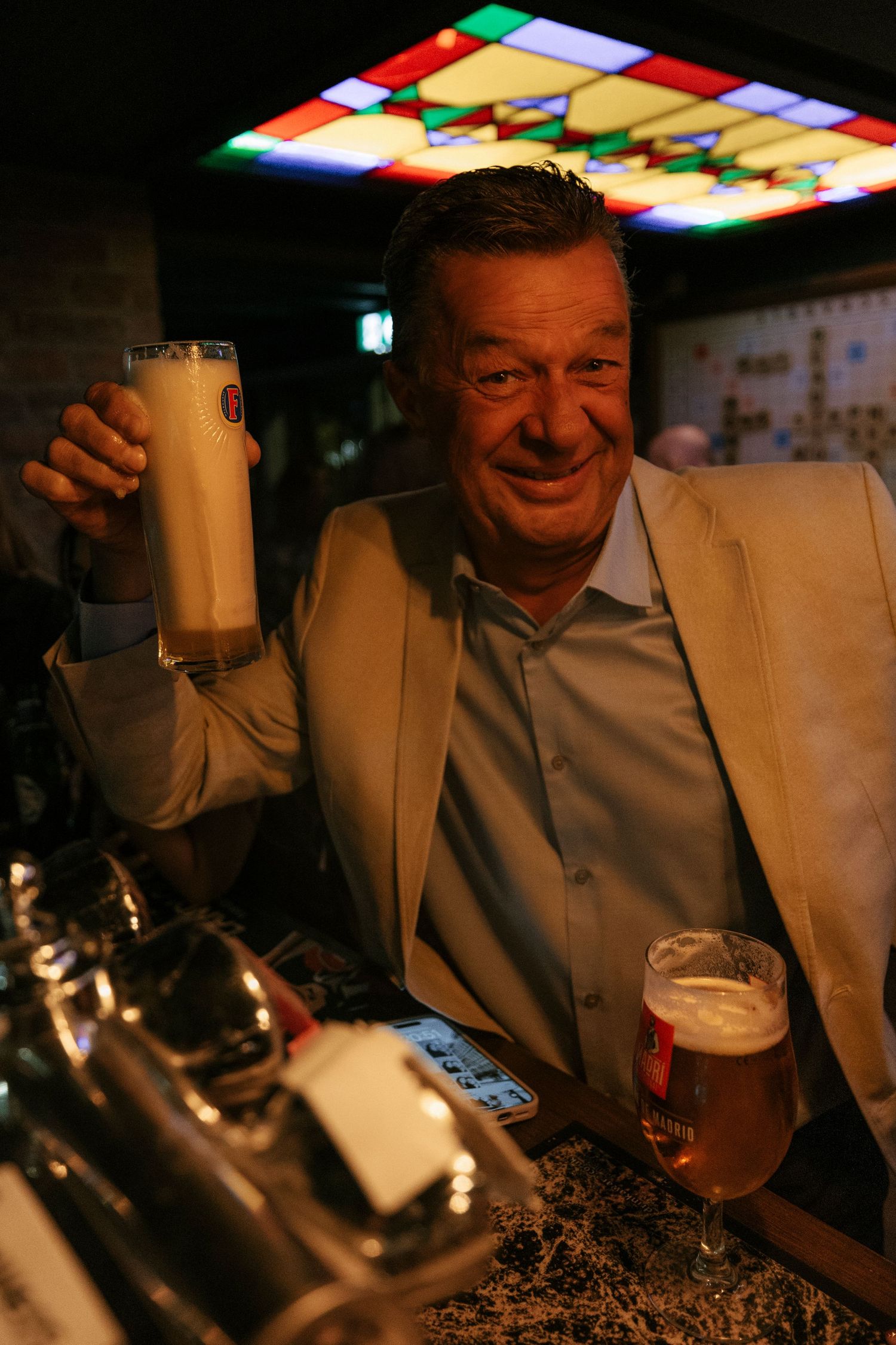 Person in beige blazer raises a large beer stein at a dimly lit bar with colorful ceiling lights.