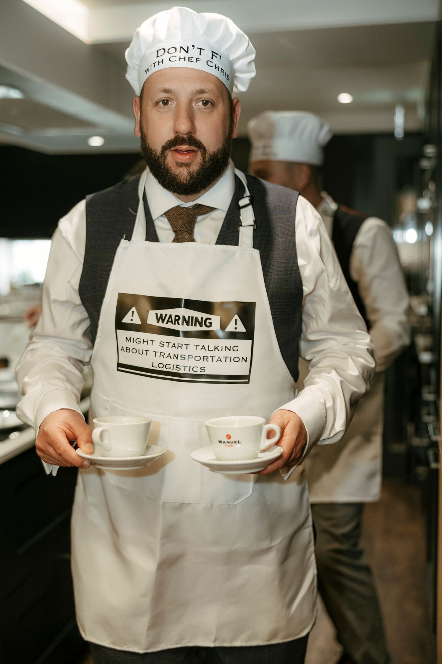Restaurant worker in chef hat and apron holds coffee cups while wearing humorous warning label on uniform.