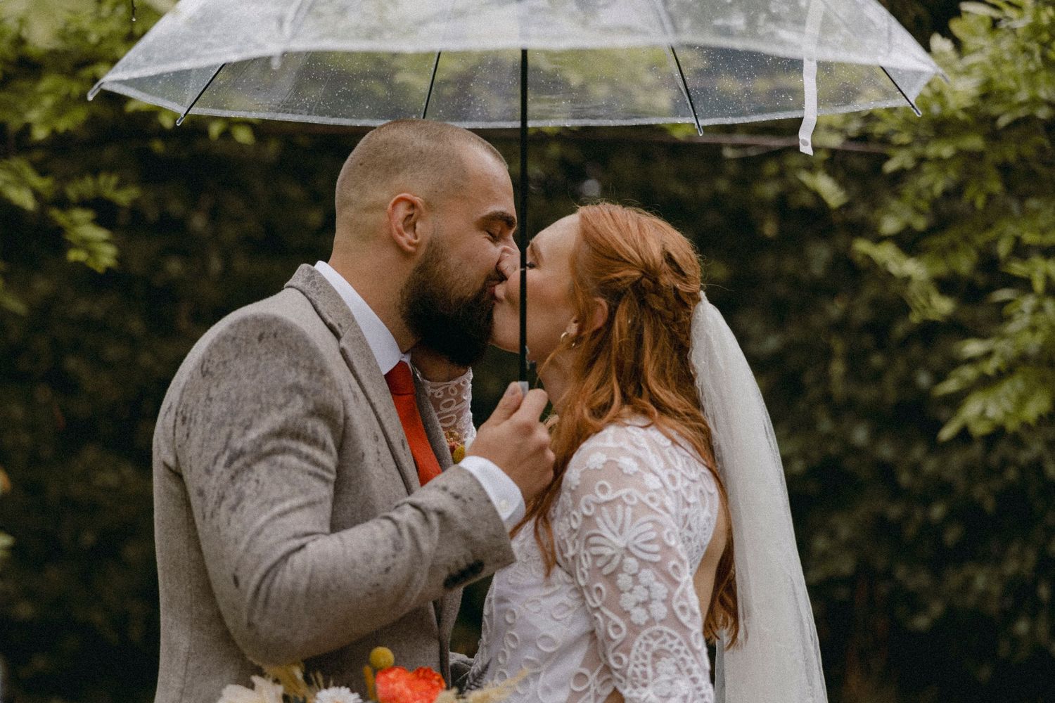 Bride and groom share a romantic kiss under a clear umbrella in the rain on their wedding day.