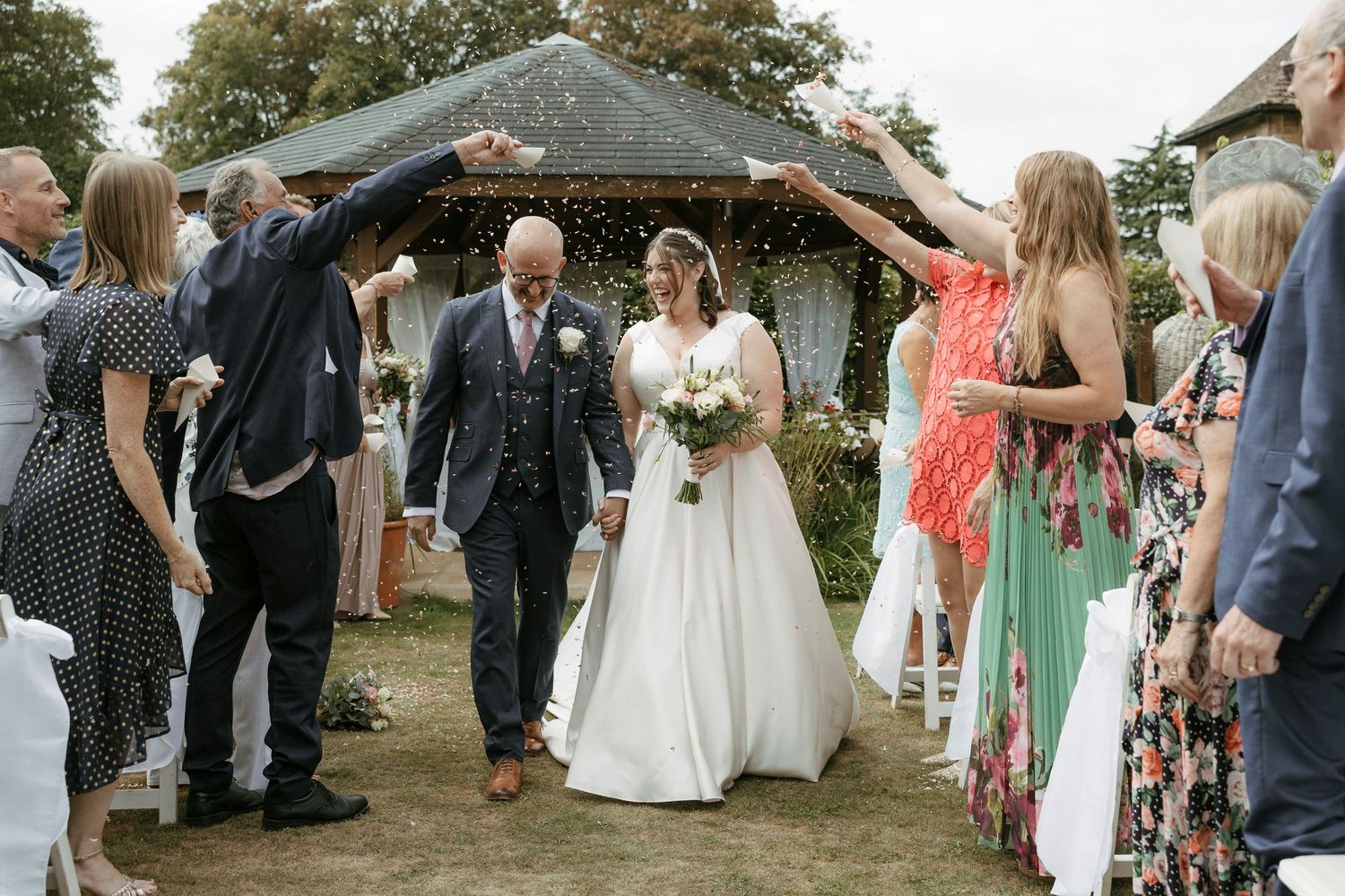 Newlyweds walk through confetti arch surrounded by cheering guests at outdoor wedding.