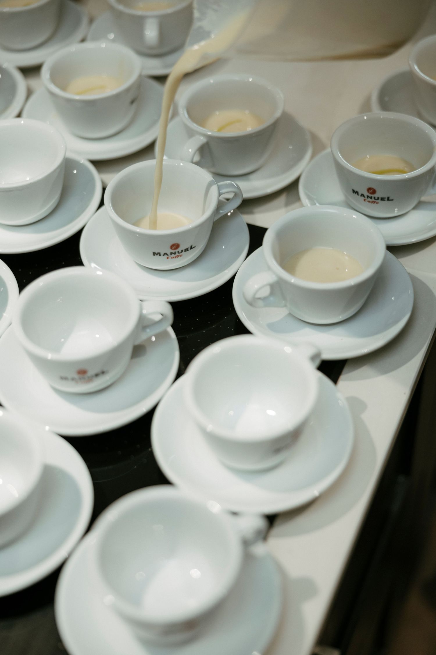 White porcelain coffee cups and saucers arranged in rows with liquid being poured into one cup.