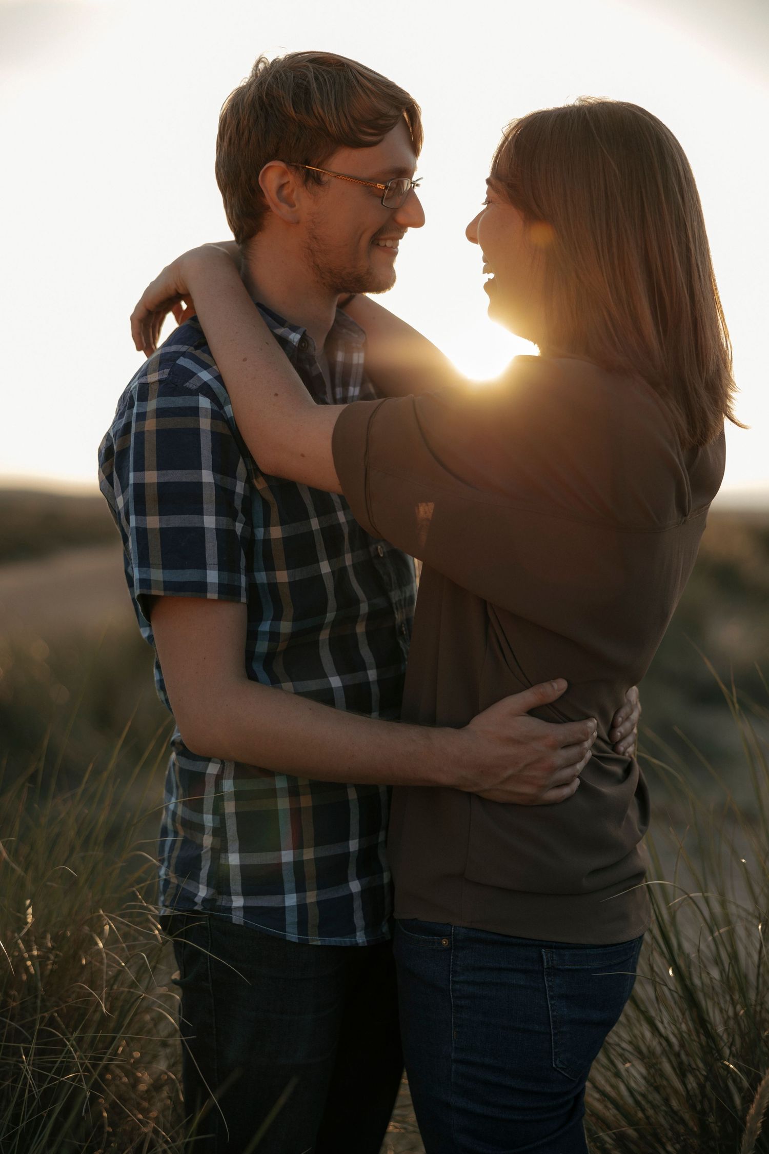 Couple embracing and smiling at sunset in a grassy field, golden backlight glowing between them.