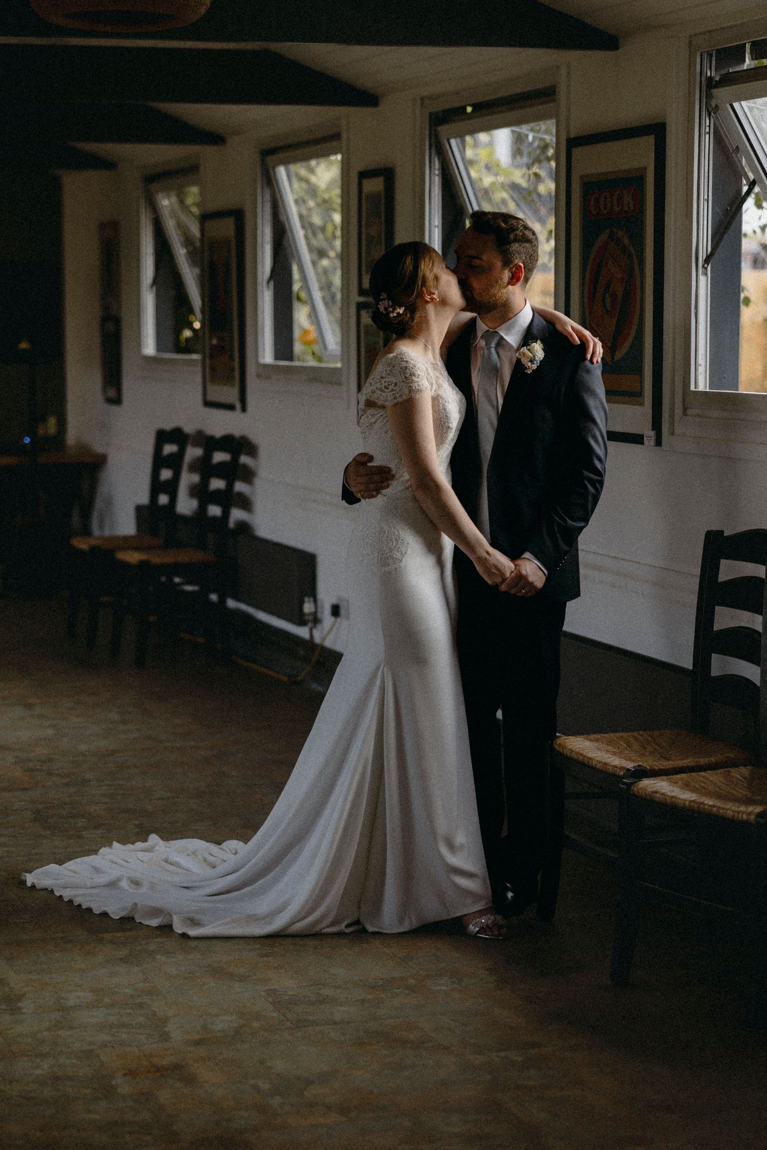A bride and groom share a romantic kiss in a rustic indoor venue, with the bride in an elegant white gown with a train.