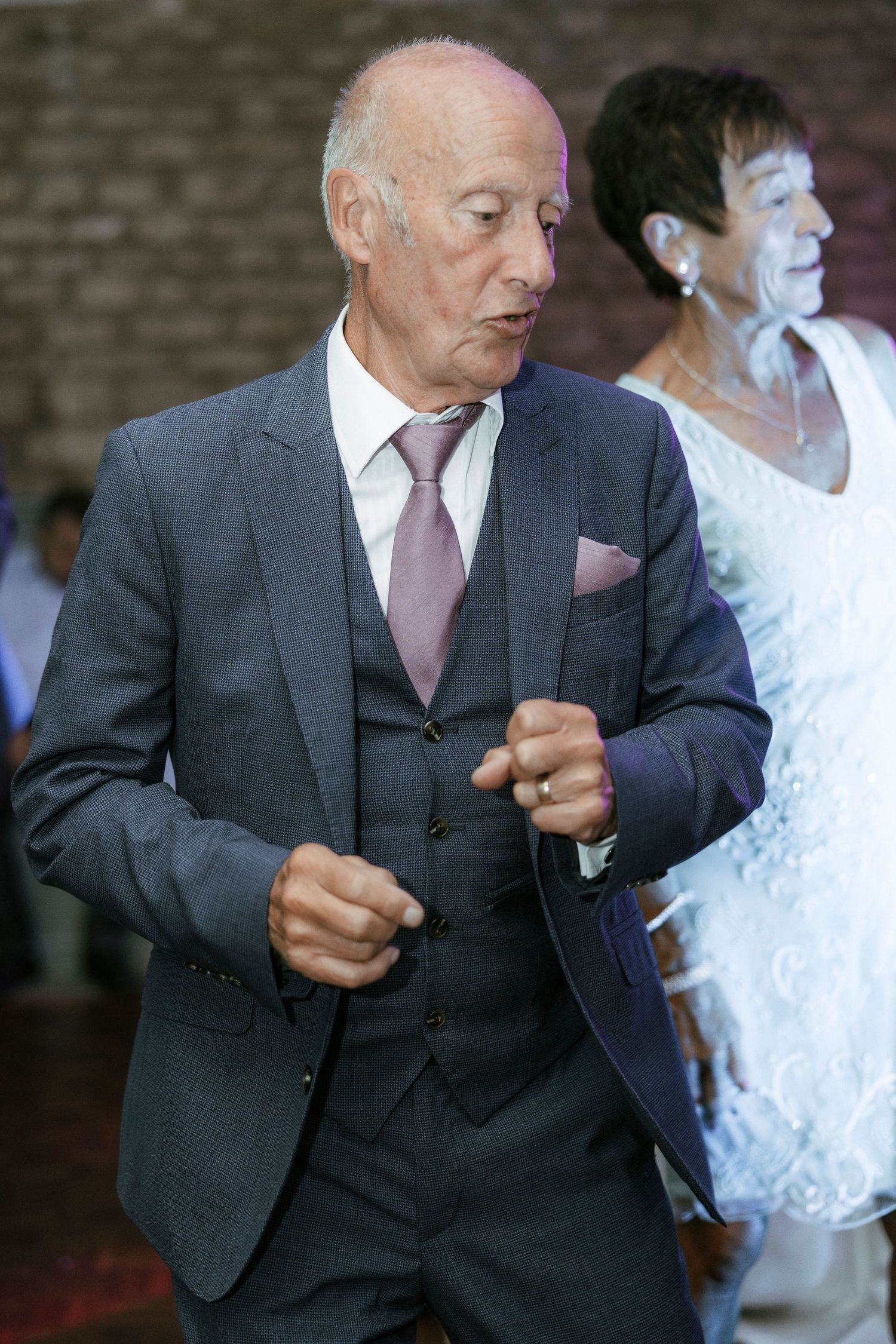 Elderly man in gray three-piece suit with pink tie dancing at a formal event, woman in white dress visible behind him.