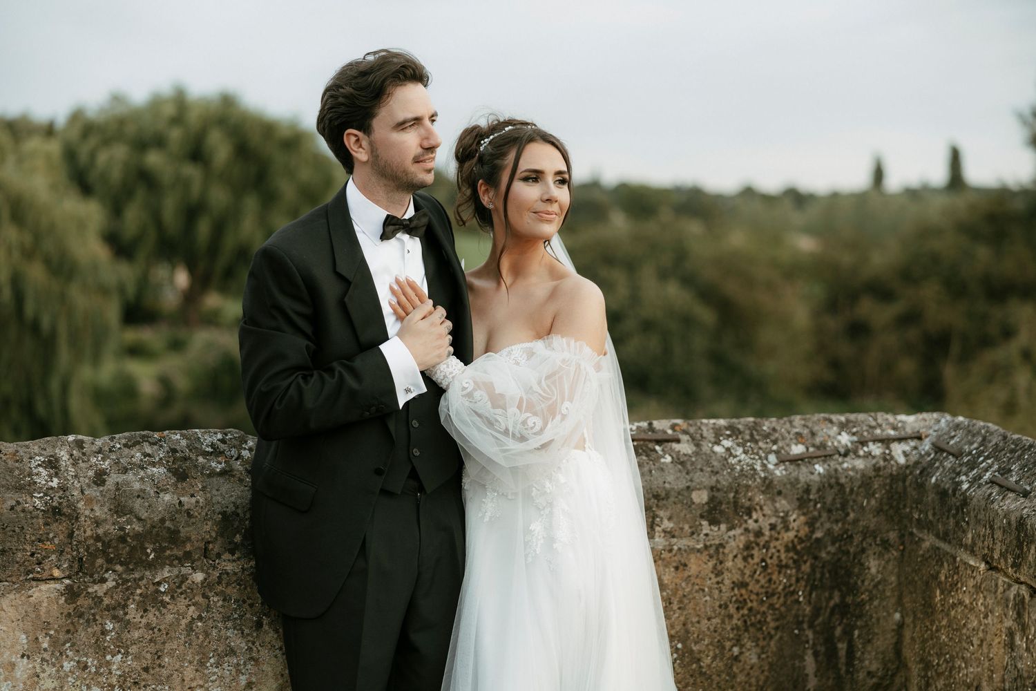 Elegant bride and groom posing outdoors on stone wall with lush greenery, groom in black tuxedo, bride in white strapless gown.