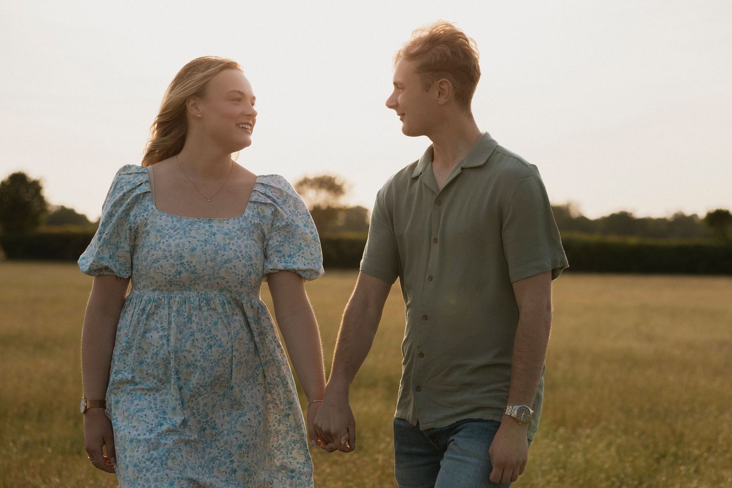 Pregnant couple holding hands smiling in golden field at sunset, wearing casual summer clothes.