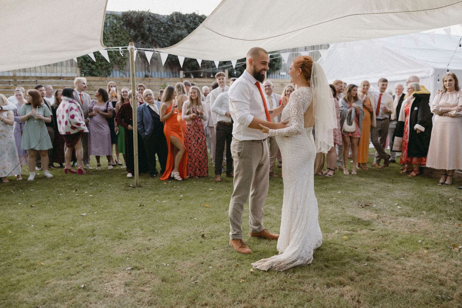 Bride and groom share first dance at outdoor wedding reception under white tent with guests watching.