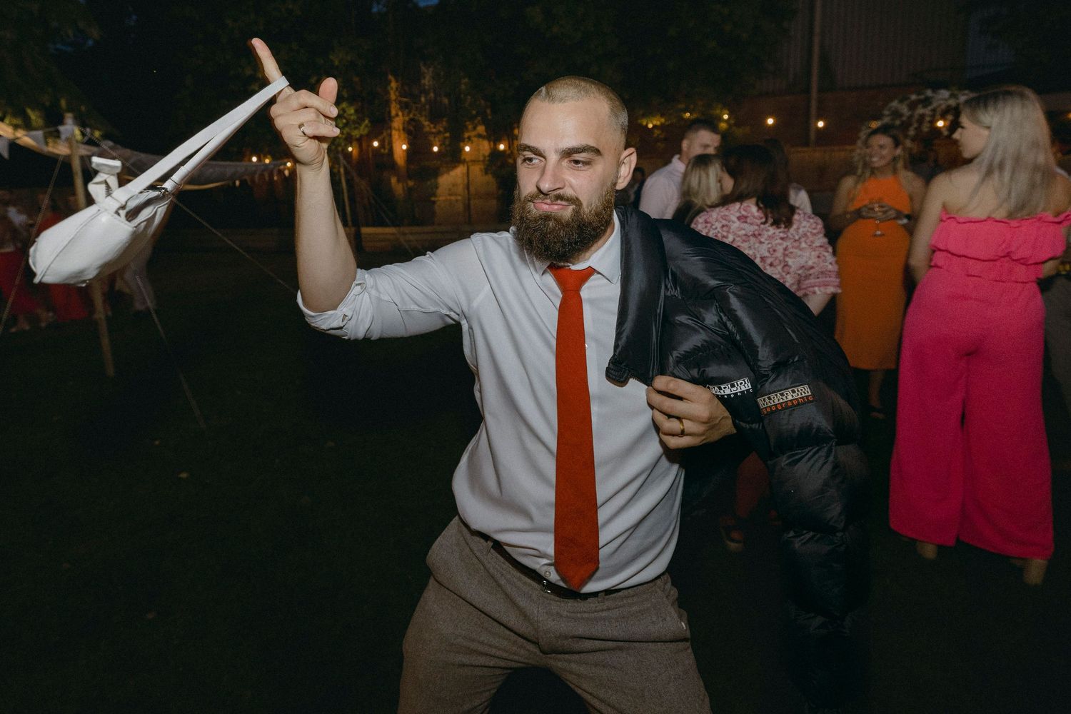 A bearded man in a white shirt and red tie dances energetically at an outdoor evening party.