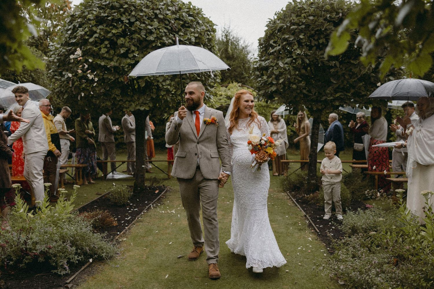 Bride and groom walk hand in hand under clear umbrellas in a garden wedding ceremony on a rainy day.
