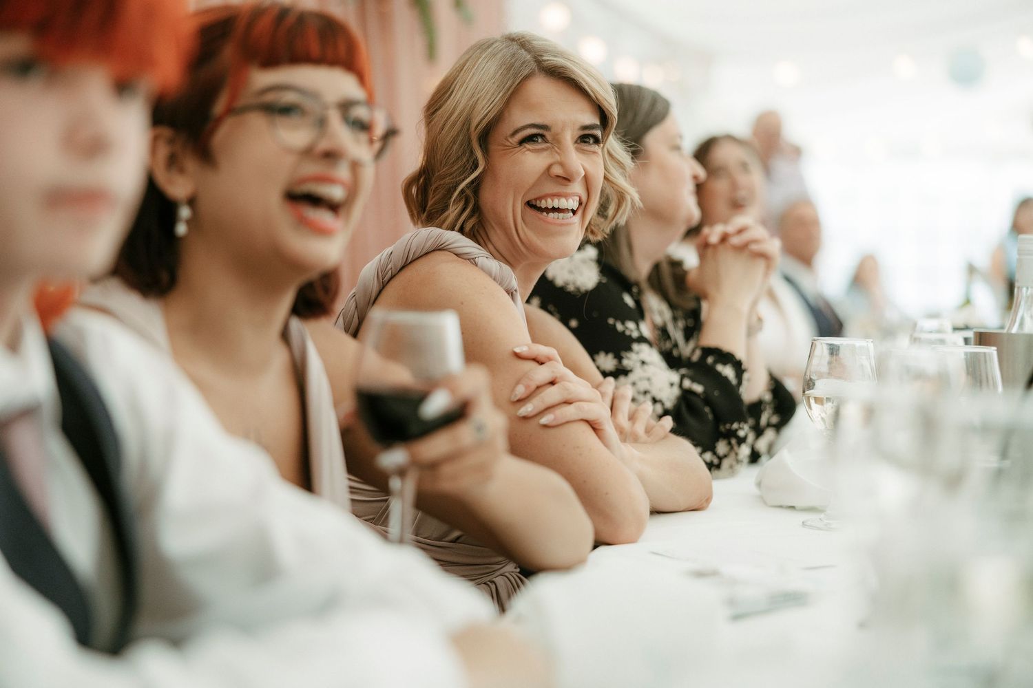 Happy wedding guests laughing and smiling at reception dinner table, woman holding glass of red wine.