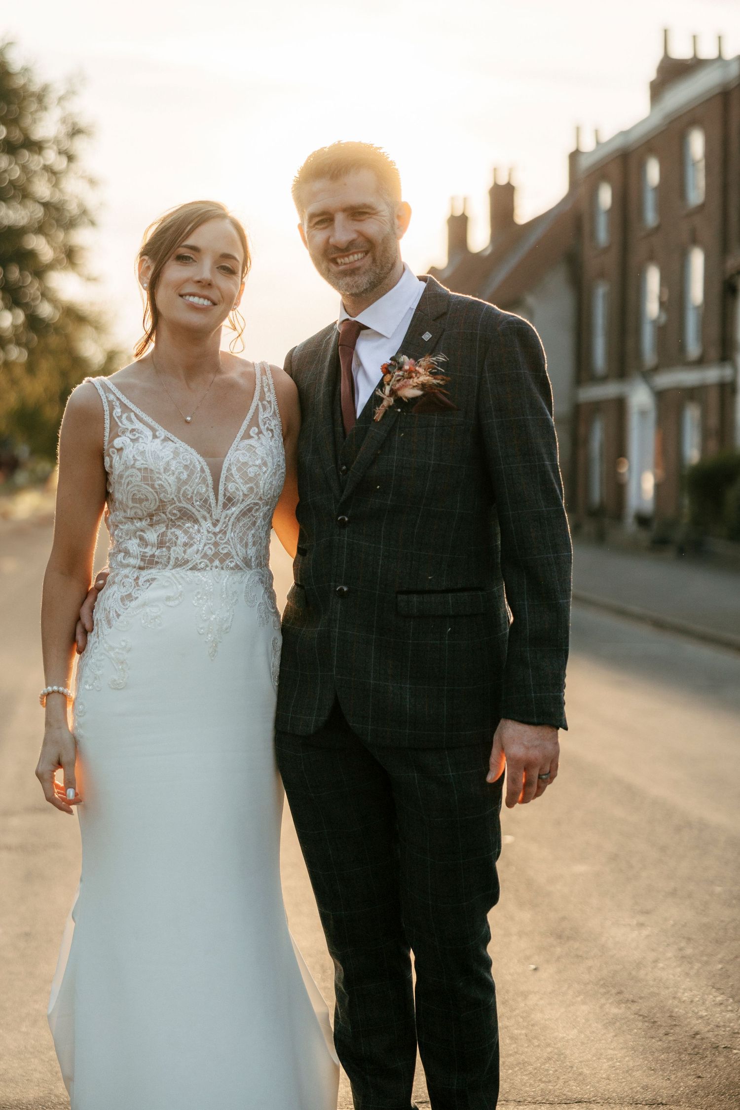 Bride in lace gown and groom in plaid suit smiling together on a street at golden hour.