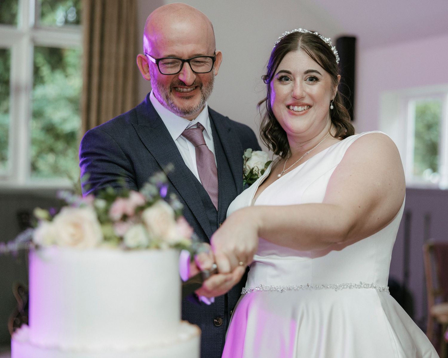 Bride and groom smiling while cutting their white tiered wedding cake at a elegant indoor wedding reception.