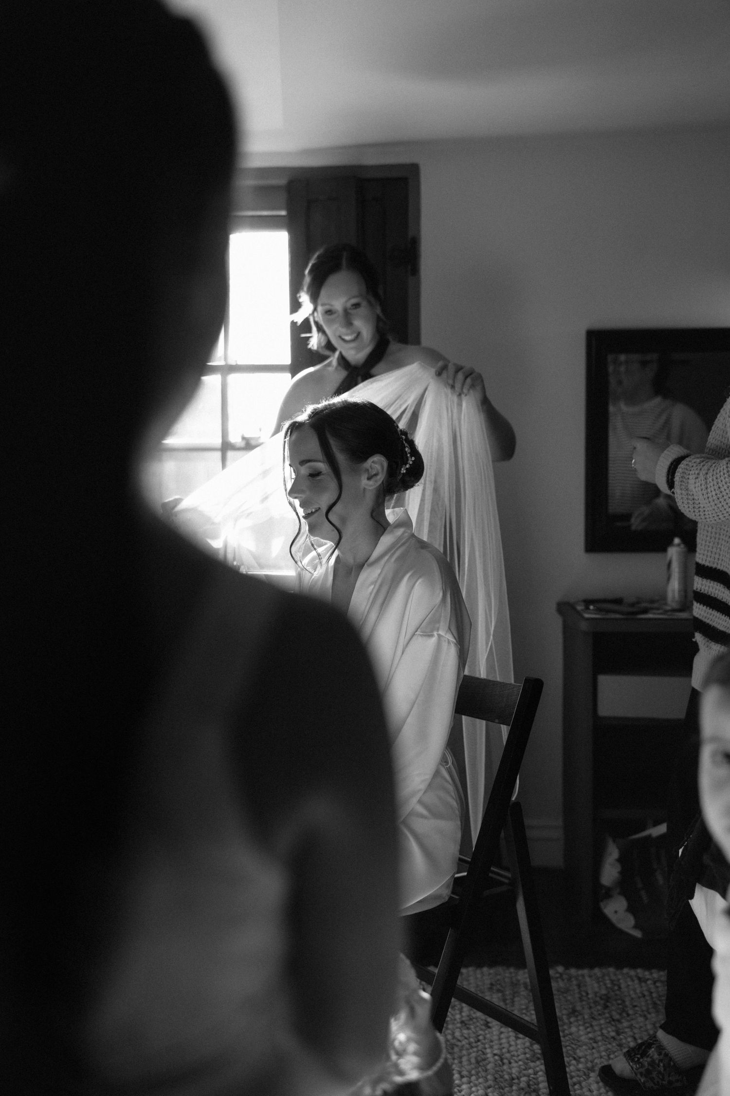 Bridesmaid helps bride with veil during wedding preparations in black and white photo.