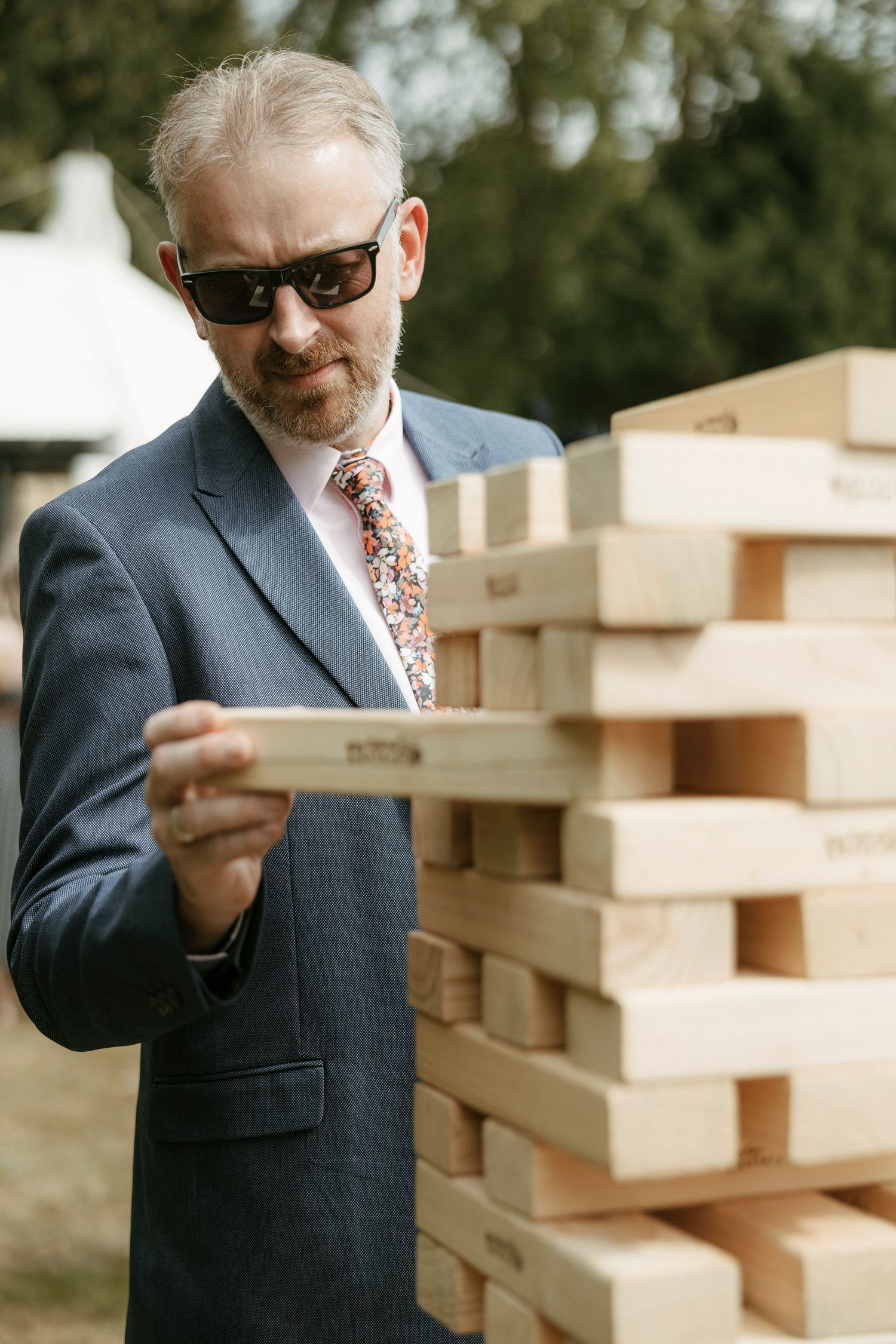 Man in suit and sunglasses playing giant Jenga at an outdoor wedding reception.