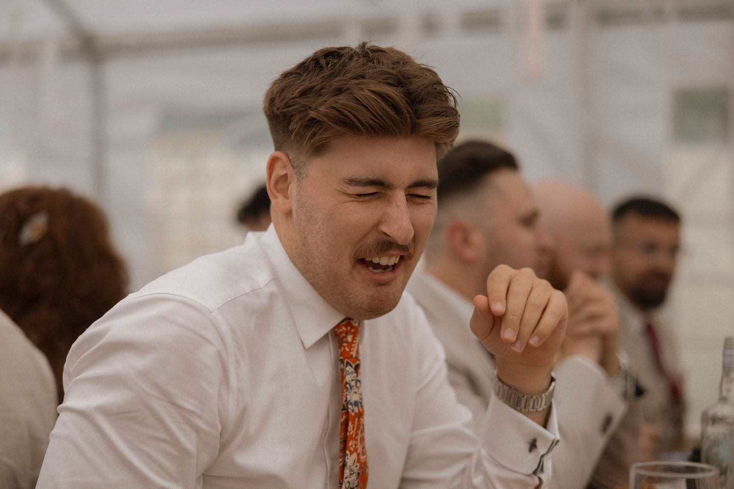 Man in white shirt and floral tie laughing joyfully at a wedding reception dinner table.