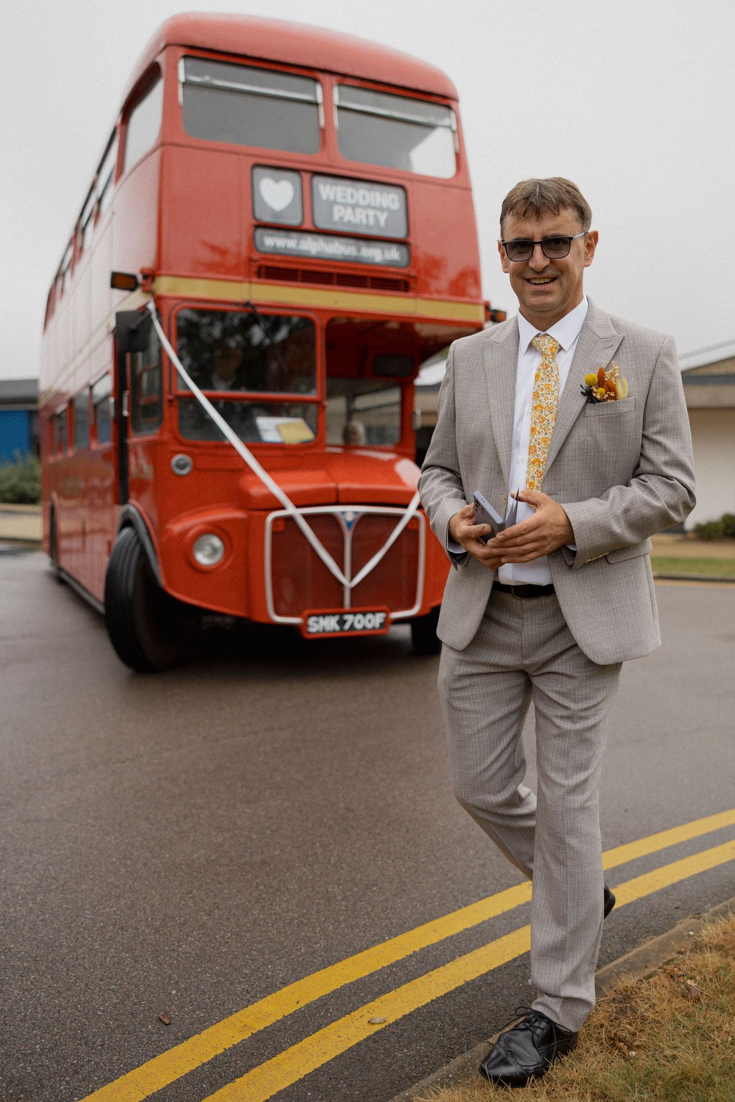 Man in gray suit with yellow tie stands before a red double-decker wedding party bus on a road.