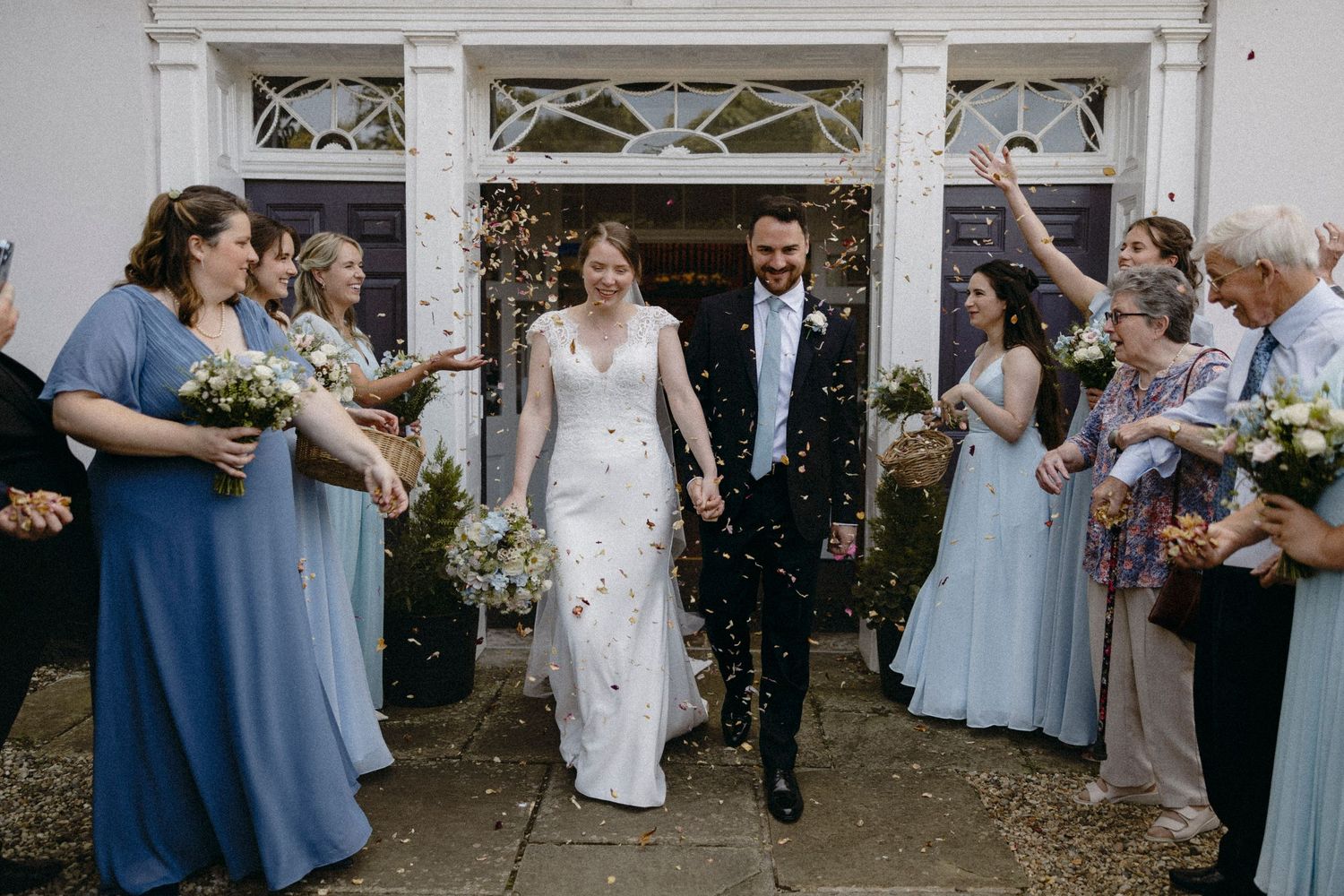Bride and groom walk through confetti shower outside white venue, flanked by bridesmaids in blue dresses and wedding guests.