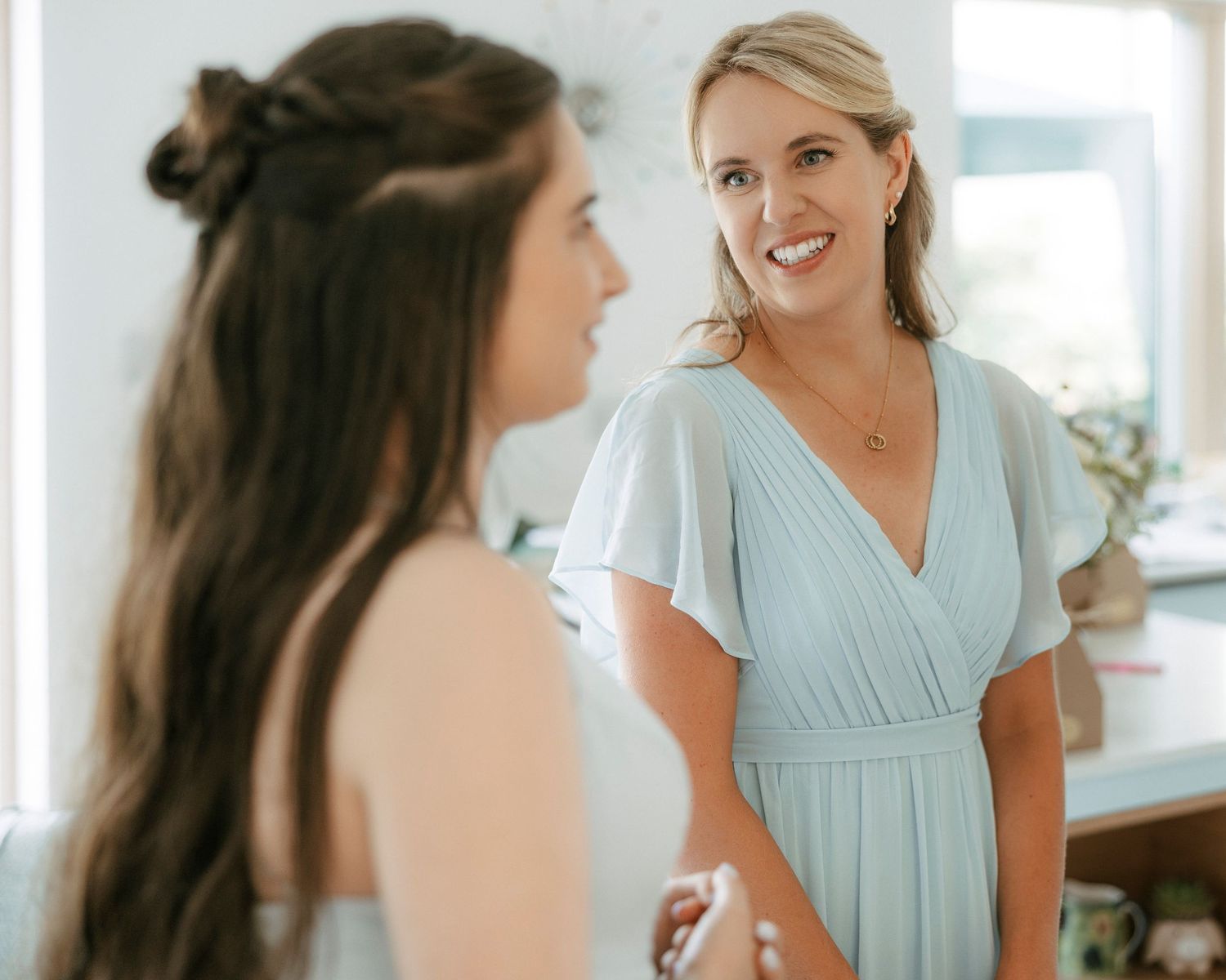 Two women in light blue dresses share a warm moment during wedding preparations in a bright, elegant room.