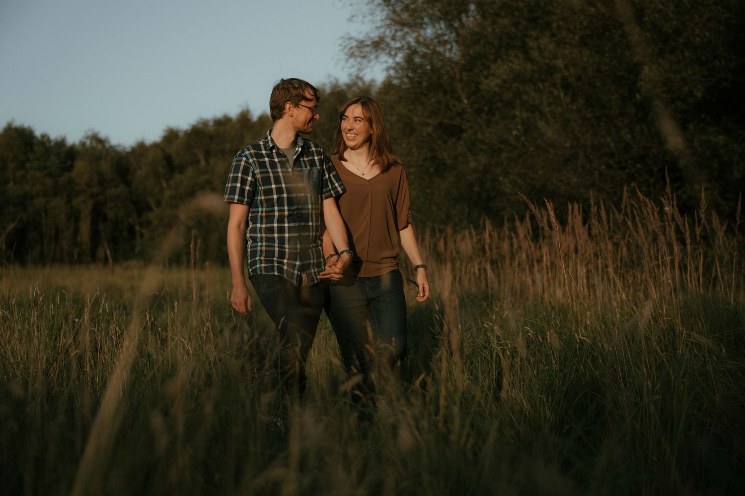 Couple walking hand in hand through a sunlit meadow with tall grass and trees in the background.