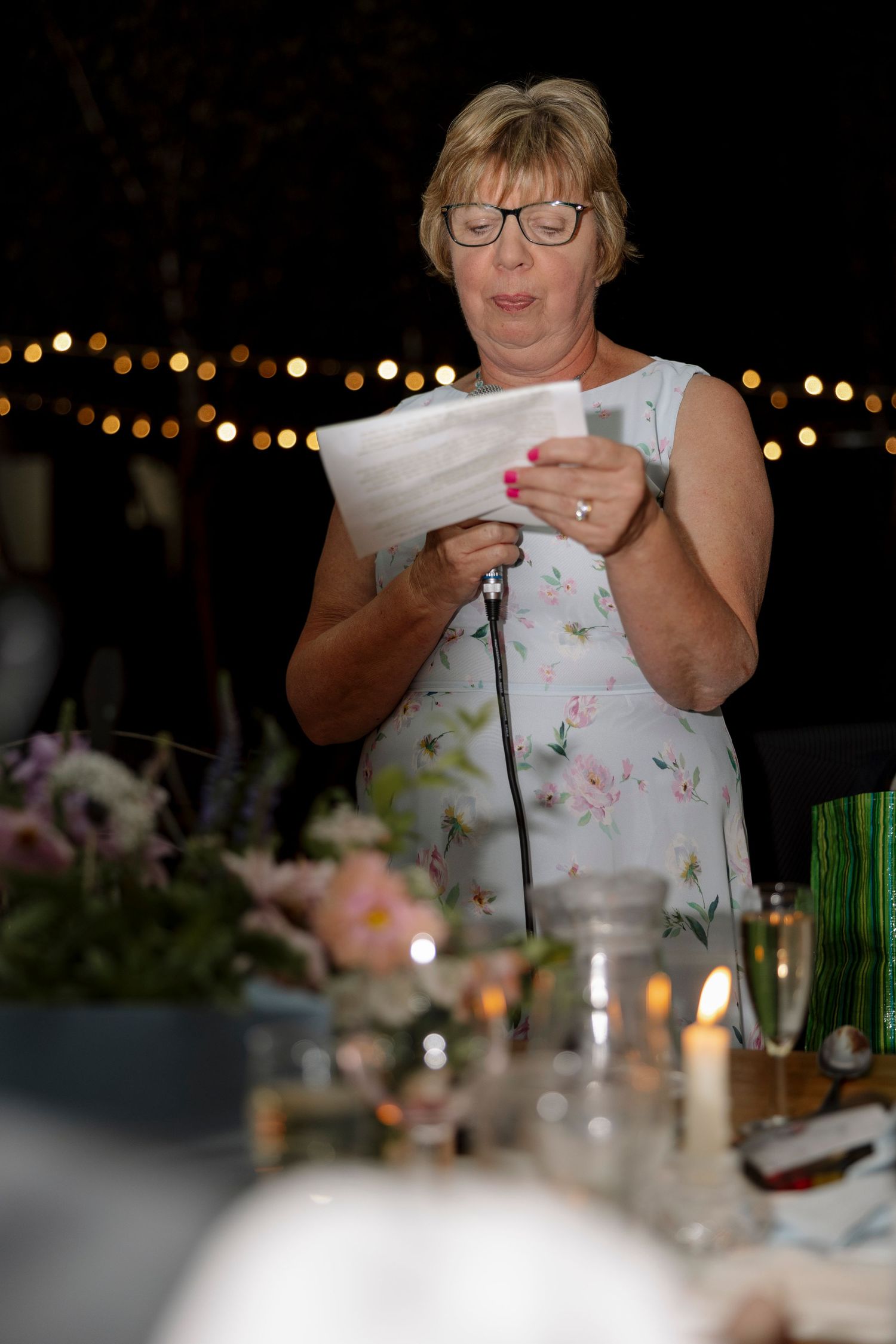 Woman reading speech at outdoor evening wedding reception with string lights and floral centerpieces.